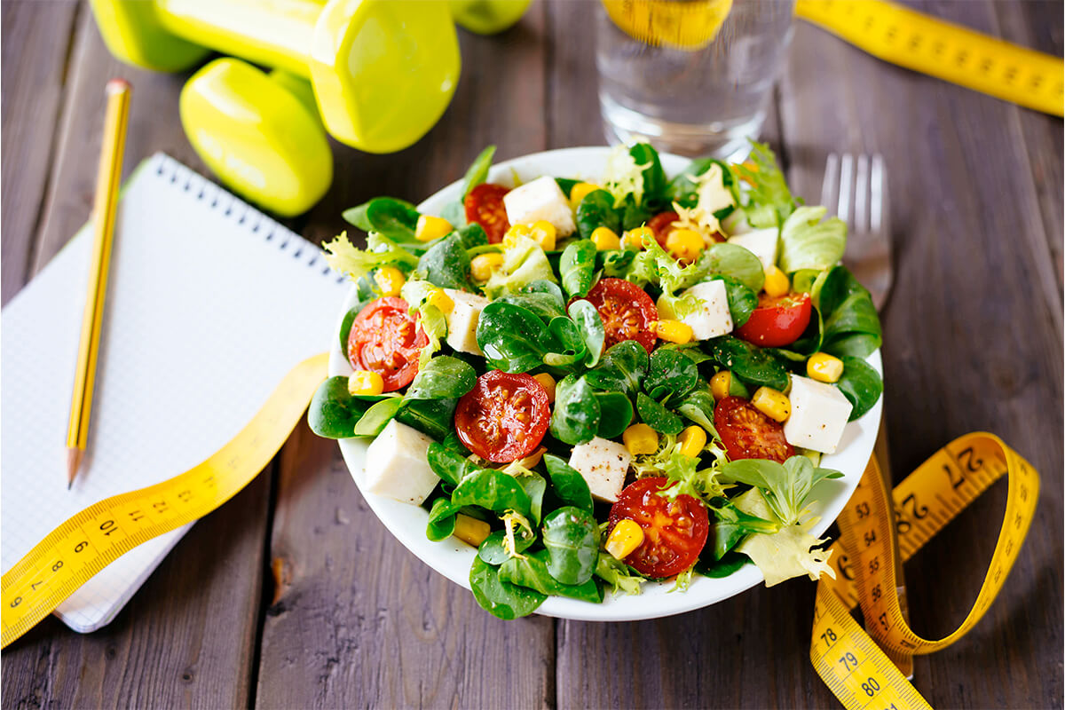 Bunte Schüssel mit frischem Fitness-Salat auf einem Holztisch, umgeben von Maßband und Notizblock.