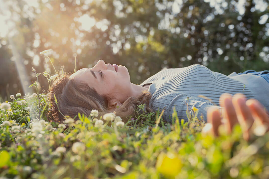 Junge Frau liegt entspannt im Gras mit geschlossenen Augen, Sonnenlicht im Hintergrund.