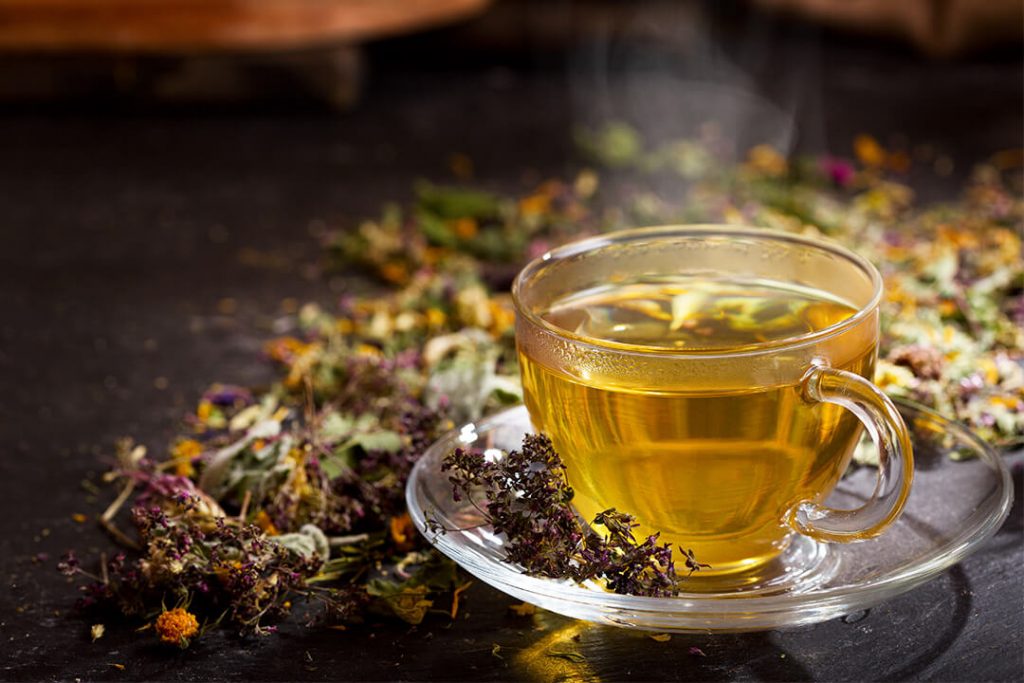 Dampfender Kräutertee in einer transparenten Tasse, umgeben von losem, getrocknetem Teekräutermix.