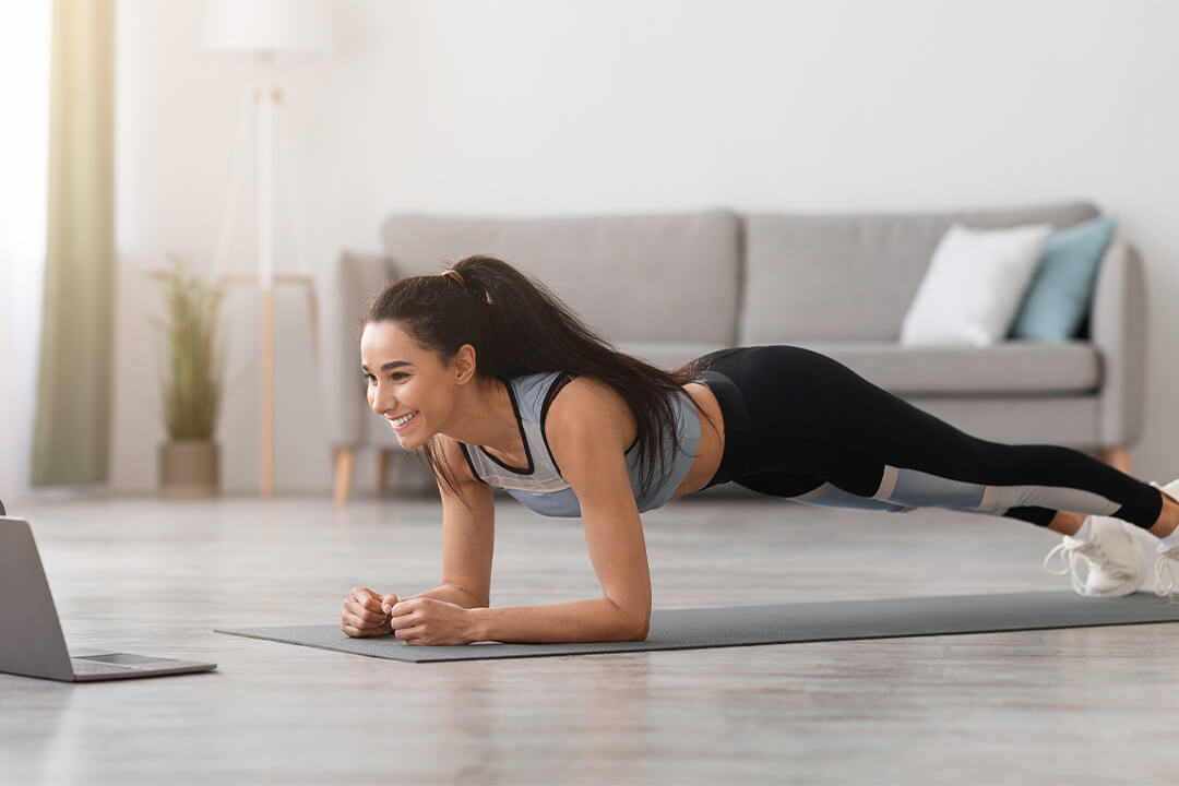 Frau mit dunklen, zusammengebundenen langen Haaren macht eine Plank-Übung auf einer Matte vor einem Laptop.